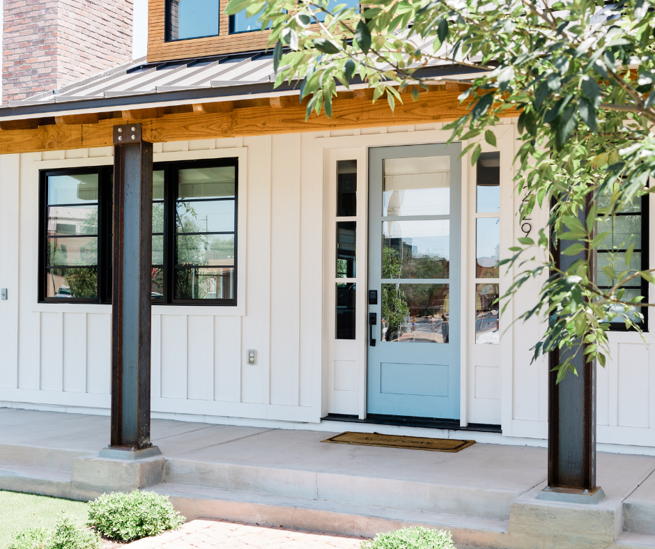 The image shows the front exterior of a modern, custom-built home. It features a light blue front door with multiple glass panels, white board and batten siding, and black-framed windows. The house has a covered porch supported by dark, metal columns and a wooden beam with visible bolts, adding a rustic touch. The area around the porch is landscaped with greenery, and there is a small doormat in front of the door. GADEG Design and Build Orange County California license #1062102