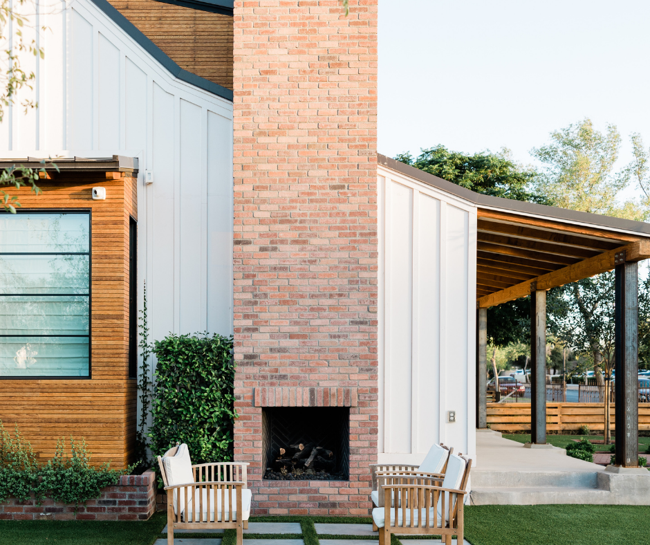 The image shows the front exterior of a modern, custom-built home. It features white vertical siding and a brick exterior fireplace, creating a striking combination of materials. The entryway includes a walnut wood door with a window, adding warmth and character to the design. The house has a covered porch supported by dark, metal columns and a wooden beam with visible bolts, enhancing the rustic touch. The area around the porch is landscaped with greenery, completing the inviting and well-crafted look of the home.