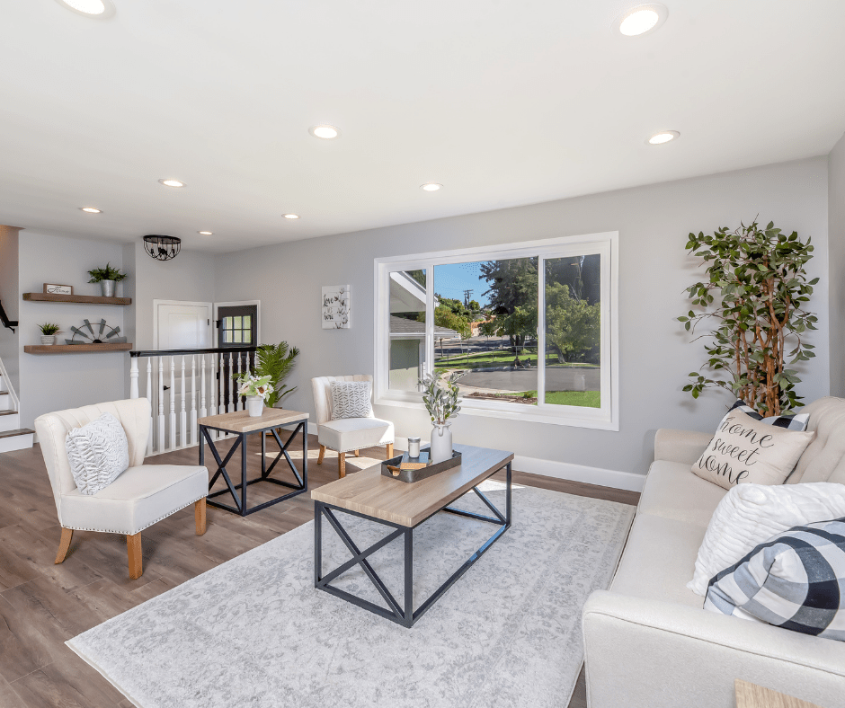 image of a grey shabby chic living room with large picture window, white railing at entry door and natural wood oak floating shelves on the wall. Orange County California GADEG Design and Build license #1062102